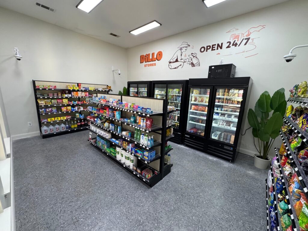 A small convenience store with shelves of snacks, drinks, and household items, plus refrigerated coolers along the back wall. A mural on the wall says Dillo Stores and Open 24/7. A large plant is near the coolers.