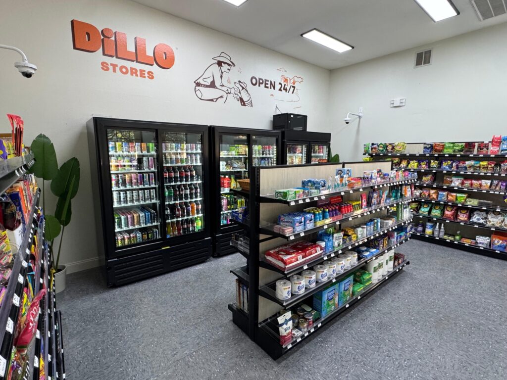 A convenience store interior with refrigerated drink coolers, shelves stocked with snacks and groceries, and a wall sign reading “DiLLO STORES OPEN 24/7” with a cowboy illustration above.