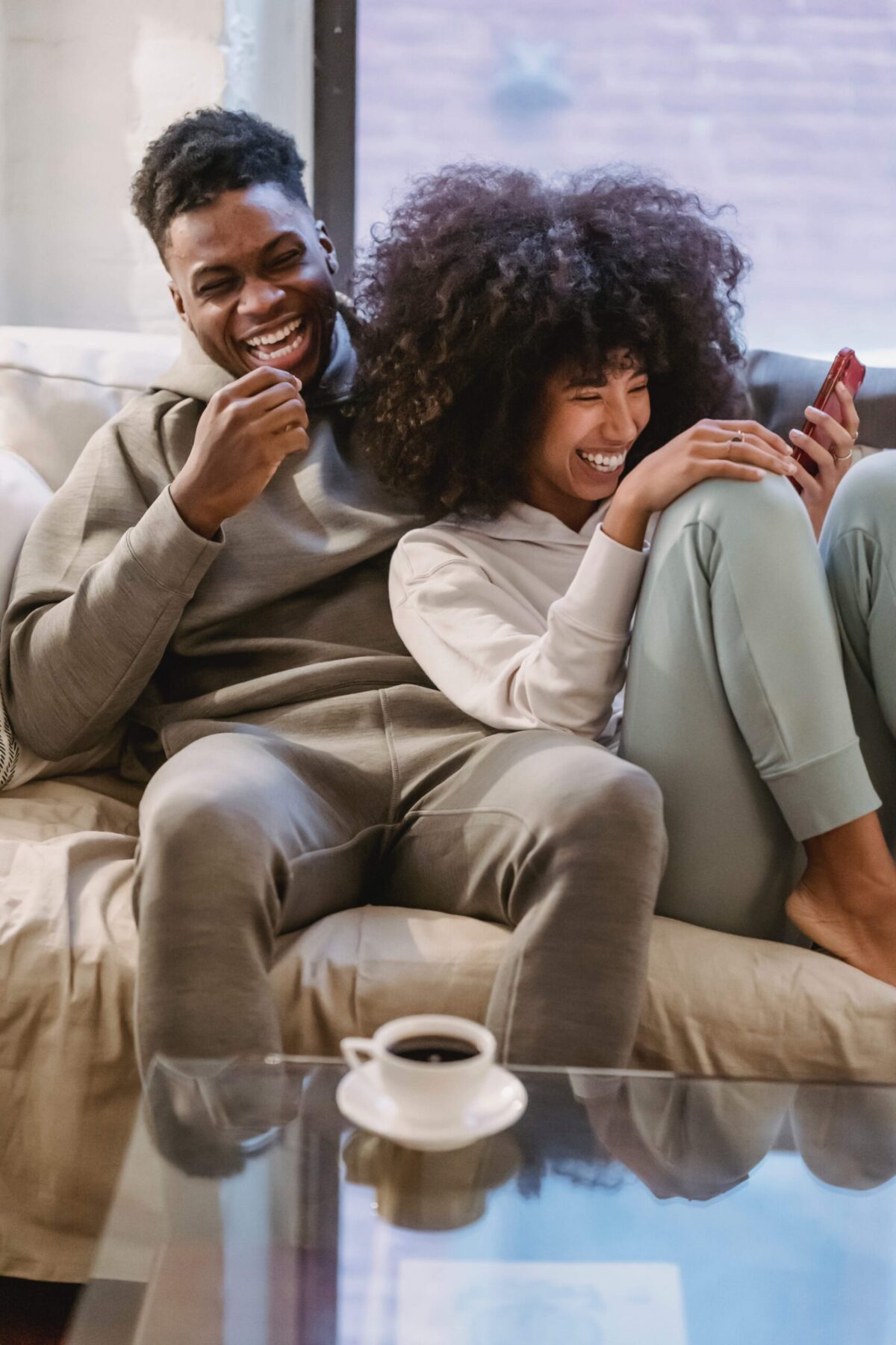 A smiling couple sits closely together on a couch, laughing while looking at a phone. The man wears a hoodie and the woman has curly hair and light-colored pants. A cup of coffee is on the glass table in front of them.